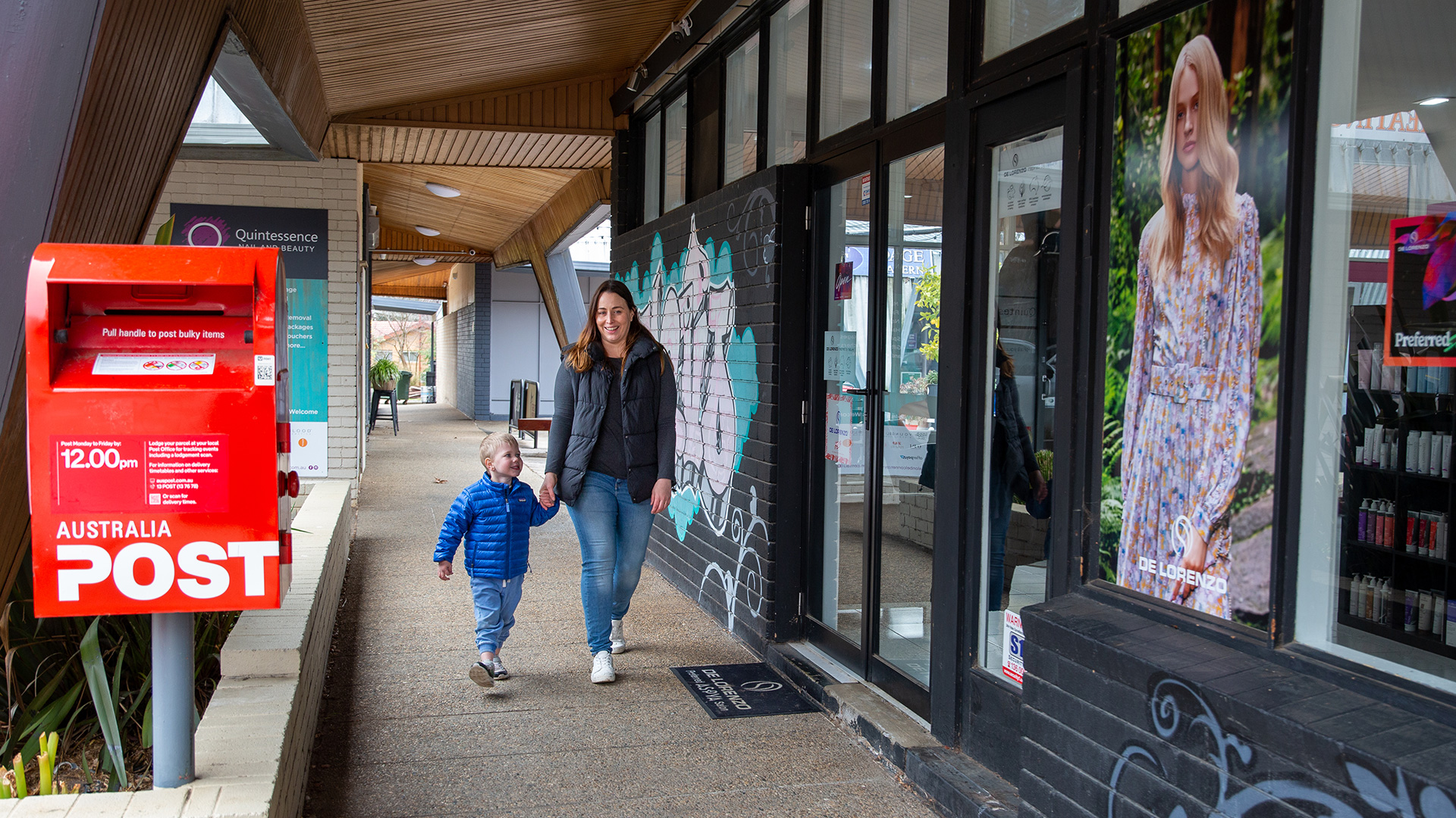 A woman and young child walk along a local shopping area next to a post box.